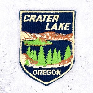Może przedstawiać: Naszywka w kształcie tarczy z ciemnoniebieskim tłem. Napis CRATER LAKE znajduje się u góry, a OREGON u dołu, oba w kolorze białym. W centrum znajduje się krajobraz z niebieskim jeziorem, zielonymi sosnami i brązowymi klifami.