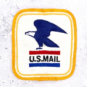 May include: A square embroidered patch with rounded corners featuring a white background and a thick yellow border. In the centre is a dark blue eagle silhouette above a red horizontal bar, the text U.S. MAIL in black, and a blue horizontal bar.