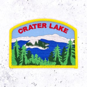 May include: A rectangular embroidered patch with a yellow border featuring a landscape of Crater Lake. The design includes a light blue sky with the red text CRATER LAKE, a deep blue lake, a central island, and green pine trees in the foreground.