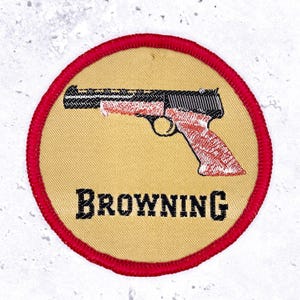 May include: A circular embroidered patch with a tan background and a red stitched border. The patch features a black and red graphic of a semi-automatic pistol and the word BROWNING in black capital letters centred below the firearm illustration.