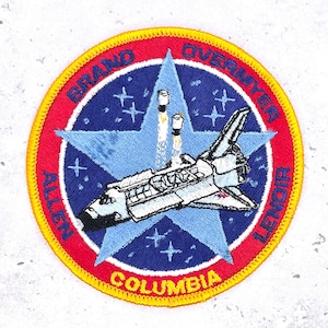 Puede incluir: Parche bordado con un diseño de transbordador espacial y la palabra "COLUMBIA" en la parte inferior. El parche tiene un borde rojo con los nombres "BRAND OVERMYER ALLEN LENDIR" alrededor de una estrella azul con un transbordador y dos cohetes.