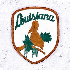 May include: A shield-shaped embroidered patch with a white background and a brown border. It features a brown pelican perched on a branch with green foliage and the word Louisiana stitched in dark green cursive script across the top.