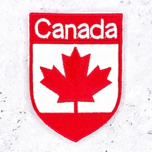 Może przedstawiać: Naszywka w kształcie tarczy z czerwoną obwódką i białym tłem. W górnej części znajduje się napis Canada białymi wielkimi literami na czerwonym polu. W dolnej części widnieje duży, szczegółowy wzór czerwonego liścia klonu.