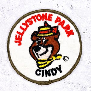 May include: Embroidered patch featuring a cartoon bear with a yellow and black scarf and hat. The patch reads "JELLYSTONE PARK" in red, with the name "CINDY" below the bear. The patch has a white background and a green border.