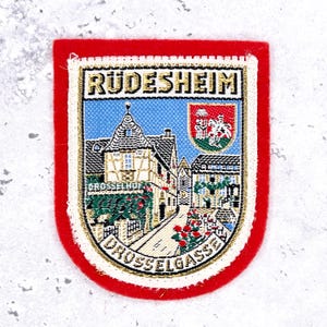 Peut inclure: Un écusson brodé rectangulaire avec une bordure en feutrine rouge représentant une scène tissée du Drosselhof à Rudesheim. Le motif inclut le texte Rudesheim et Drosselgasse, un ciel bleu, un petit blason et une vue détaillée d'une maison à colombages.