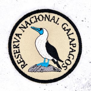 May include: A circular patch featuring a blue-footed booby bird illustration. The bird is white with black wings and a blue beak and feet, perched on a gray rock. The text "RESERVA NACIONAL GALAPAGOS" surrounds the bird in a black border.