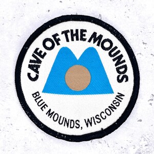 May include: Circular patch with the text "CAVE OF THE MOUNDS BLUE MOUNDS, WISCONSIN". The patch features a blue mountain design with a tan circle in the center, all set against a white background with a black border.