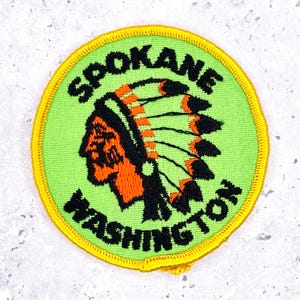 May include: Embroidered patch with the words "Spokane Washington" in black lettering around the edge. The patch features a Native American headdress in orange, black, and green. The patch has a yellow border.