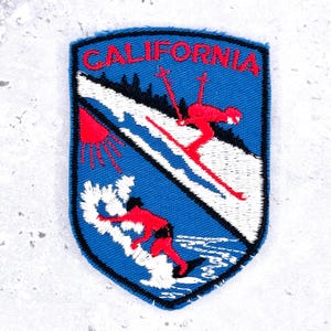 May include: A shield-shaped embroidered patch with a blue background featuring the word CALIFORNIA in red text. The design depicts a red skier on a white snowy slope above a red surfer riding a white wave, with a red sun on the left side.