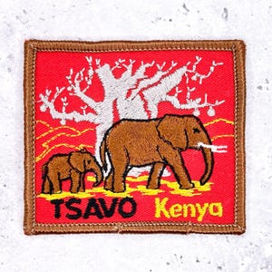 May include: Square patch with a red background featuring two brown elephants, a white tree, and yellow accents. The text "TSAVO Kenya" is displayed in yellow and black. The patch has a brown border.