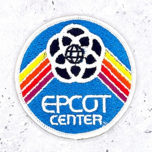 May include: A circular embroidered patch featuring a blue background with a white, black, and blue globe icon at the top. Three diagonal stripes in yellow, orange, and red extend from the globe. The text EPCOT CENTER is stitched in white.