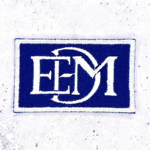 May include: A rectangular embroidered patch featuring a deep blue background with a white border. The center displays the white stitched letters E, E, and M, with the letter M partially enclosed by a curved line extending from the letter E.