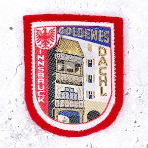 May include: A rectangular embroidered patch with a red felt border featuring the Goldenes Dachl building in Innsbruck. The design includes a red eagle emblem, the text INNSBRUCK vertically, and GOLDENES DACHL horizontally in gold thread.