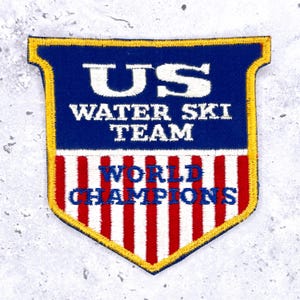 May include: Embroidered patch featuring the text "US WATER SKI TEAM WORLD CHAMPIONS." The patch has a shield shape with a blue top, red and white stripes, and a gold border. A vintage sports collectible.