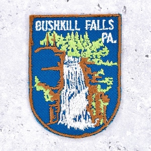 Peut inclure: Écusson brodé représentant une cascade avec le texte "Bushkill Falls PA" en blanc sur fond bleu. L'écusson a une bordure marron et représente des arbres verts et bruns.