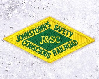 May include: Embroidered patch in a diamond shape with yellow and green colors. The patch reads "Johnstown's Safety Conscious Railroad" with "J&SC" in the center. The patch is likely for a railroad worker's uniform.