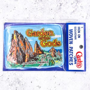 Op de afbeelding: Een rechthoekige geborduurde patch met een landschap van Garden of the Gods in Colorado Springs met oranje en grijze rotsformaties, groene bomen en een blauwe lucht. De tekst luidt Garden of the Gods en Colorado Springs in verpakking.
