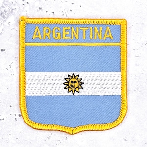 May include: A rectangular embroidered patch featuring the flag of Argentina with light blue and white horizontal stripes and a central yellow sun emblem. The word ARGENTINA is stitched in yellow capital letters at the top, all bordered by a thick yellow thread edge.