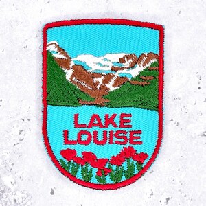 May include: A shield-shaped embroidered patch with a red border. The design features a light blue sky, brown mountains with white snow, green hills, and the red text LAKE LOUISE above a row of red flowers with green stems on a light blue base.
