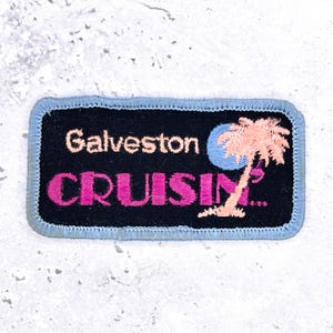 Può includere: Una toppa ricamata rettangolare con sfondo nero e bordo cucito azzurro. Presenta la scritta Galveston in bianco e CRUISIN... in rosa acceso, accompagnata da un cerchio azzurro e una palma color pesca.