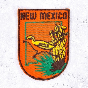 Op de afbeelding: Geborduurde patch met de tekst "NEW MEXICO" in bruin. De patch heeft een oranje achtergrond met een groene rand en toont een roadrunner en woestijnplanten in geel, groen en bruin.