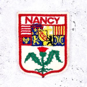 May include: Embroidered patch featuring the word "NANCY" in red letters at the top. The patch has a white background with a red border and a detailed emblem with a thistle at the bottom. The emblem includes various heraldic symbols.