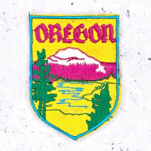 Könnte beinhalten: Gestickter Aufnäher mit gelbem Hintergrund und Schildform. Der Aufnäher zeigt das Wort "OREGON" in Pink, eine Bergkette, Bäume und einen Fluss. Der Rand ist türkis.