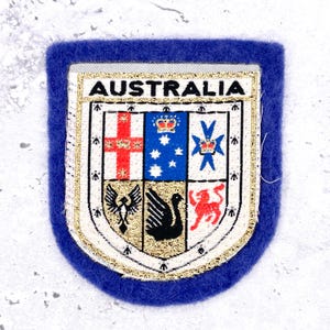 May include: A blue felt patch featuring the Australian coat of arms embroidered in white, gold, red, and black thread. The shield displays six sections with symbols including a cross, stars, a crown, a bird, a swan, and a lion, with AUSTRALIA at the top.