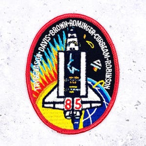 May include: Oval-shaped embroidered patch with a red border. The patch features a space shuttle design with the number "85" at the bottom. The design includes a yellow and orange sun, a blue and white planet, and the names of the crew members.