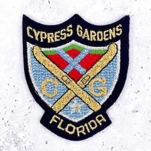 Può includere: Toppa ricamata a forma di scudo blu navy. La toppa presenta le parole "CYPRESS GARDENS FLORIDA" in filo d'oro. All'interno dello scudo ci sono sci dorati incrociati, un disegno rosso e verde e le lettere "C" e "G".