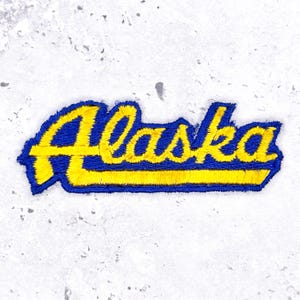Può includere: Una toppa ricamata che riporta la parola Alaska in un carattere corsivo stilizzato. Le lettere sono di colore giallo brillante con un bordo spesso blu scuro e una linea orizzontale gialla che parte dalla base della A maiuscola.