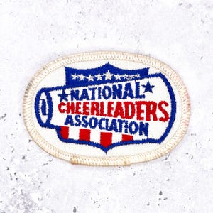 May include: Oval-shaped patch with the text "NATIONAL CHEERLEADERS ASSOCIATION" in red and blue. The patch has a white background with a blue and red design at the top and bottom. The patch is bordered with a white stitched edge.