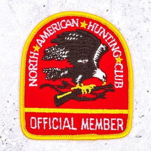 May include: Embroidered patch with a red background and a yellow border. The patch features a brown and white eagle perched on a branch, with the text "NORTH AMERICAN HUNTING CLUB" and "OFFICIAL MEMBER" in white.