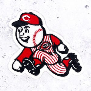 May include: An embroidered patch featuring a cartoon baseball character with a white face, red cap with a white C, and a red and white pinstriped uniform. The character is in a running pose, wearing black cleats and red sleeves, with a red C logo.