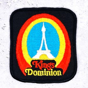 Peut inclure: Écusson brodé avec une bordure noire. Il présente un dessin de tour blanche à l'intérieur d'un cercle bleu, jaune et rouge. Les mots "Kings Dominion" sont en écriture jaune et rouge en dessous.