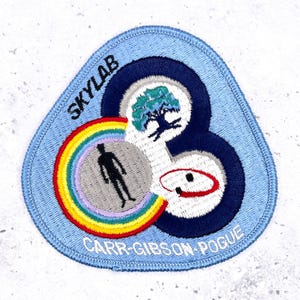 May include: Embroidered Skylab mission patch with a light blue border. The patch features a rainbow, a silhouetted figure, a tree, and the text "SKYLAB" and "CARR-GIBSON-POGUE". The design is a mix of colors including blue, green, red, and yellow.