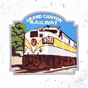 May include: Embroidered patch featuring a train with the text "Grand Canyon Railway." The train is white and yellow with brown accents, set against a blue sky. The patch includes the number "6703."