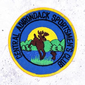 Pode incluir: Emblema bordado circular com o texto "CENTRAL ADIRONDACK SPORTSMEN'S CLUB". Apresenta um alce castanho, árvores verdes e um sol amarelo sobre um fundo azul, com uma borda amarela.