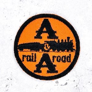 May include: A circular embroidered patch featuring a vibrant orange background with a thick black border. The design includes a black silhouette of a steam locomotive train in the center, with the black text A & A railroad displayed prominently.
