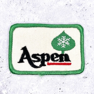 Könnte beinhalten: Ein rechteckiger Aufnäher mit grünem Rand und weißem Hintergrund. Das Wort "Aspen" ist in Schwarz gestickt, darunter eine rote Linie. Rechts ist ein grüner Baum mit einer weißen Schneeflocke.