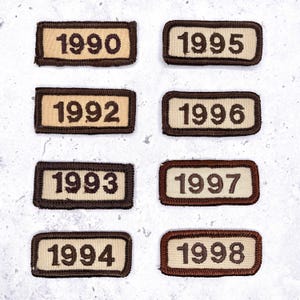 May include: Eight rectangular patches with the years 1990, 1992, 1993, 1994, 1995, 1996, 1997, and 1998 embroidered on them. Each patch has a beige background with brown text and a brown border. The patches are arranged in two rows of four.