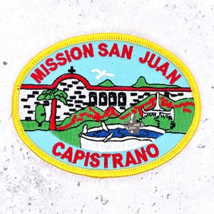 May include: An oval embroidered patch with a yellow border featuring a light blue sky, a white mission building with a red arch, a green palm tree, and a fountain. The text MISSION SAN JUAN CAPISTRANO is stitched in red capital letters.