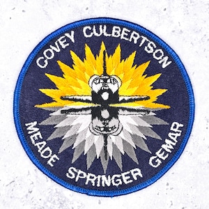 May include: Embroidered patch with a circular design featuring a central spacecraft image with outstretched wings, surrounded by a yellow and white burst. The patch has a navy blue background with white text around the edge, including names.