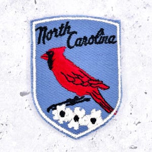 Peut inclure: Un écusson brodé en forme de bouclier avec un fond bleu clair et une bordure blanche. Il représente un cardinal rouge vif perché sur des fleurs de cornouiller blanches à cœur noir. Le texte North Carolina est brodé en écriture noire.