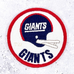 May include: A circular embroidered patch featuring a dark blue football helmet with a white face mask. The word GIANTS is stitched in white across the helmet and in dark blue below it. The patch has a red border and includes the letters TM.