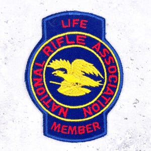 Könnte beinhalten: Gestickter Aufnäher mit blauem Hintergrund und gelbem und rotem Text. Der Aufnäher trägt oben "LIFE", "NATIONAL RIFLE ASSOCIATION" um einen gelben Adler herum und unten "MEMBER". Der Aufnäher hat eine Schildform.