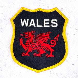 May include: A shield-shaped patch with a black background and a yellow border. The word "WALES" is in white block letters at the top. A red Welsh dragon is embroidered in the center.