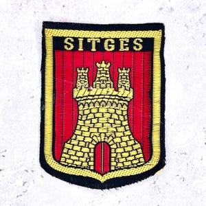 Peut inclure: Un écusson brodé rectangulaire présentant un fond rouge avec un motif de château jaune et le mot SITGES en lettres capitales jaunes en haut. L'écusson est bordé par un contour noir épais et un cadre intérieur jaune fin.