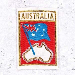 May include: A rectangular embroidered patch featuring the word AUSTRALIA in gold thread at the top. Below, a blue Australian flag with white stars is depicted over a white outline of the Australian continent on a red background with gold trim.