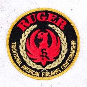May include: A circular embroidered patch with a black background and a gold border. It features a red Ruger phoenix logo in the center, surrounded by a gold laurel wreath. The text RUGER appears in red at the top, with TRADITIONAL AMERICAN FIREARMS CRAFTSMANSHIP in gold at the bottom.